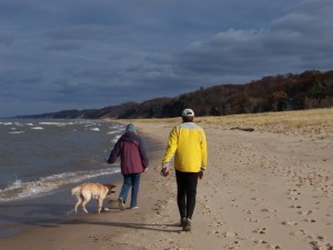 Color photograph of the shore of Lake Michigan taken by Anna J. Cook in late October at the Saugatuck Dunes State Park in Michigan in midmorning. On the left is the lake with small waves coming in. In the center are Addie (the golden retriever puppy), Janet and Mark (Anna's parents), all walking away from the camera down the beach. On the right are grass-covered dunes and in the background are late-autumn trees. The sky is partially overcast.