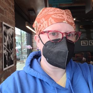 A self portrait wearing a headscarf, blue hoodie, red-rimmed glasses, and a black kn95 filtration mask.